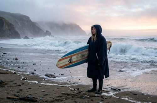 Surfer wearing a hooded warm surf poncho on a chilly ocean beach.