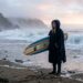 Surfer wearing a hooded warm surf poncho on a chilly ocean beach.
